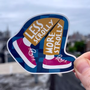 May include: A sticker with a blue and white plaid background featuring a cartoon illustration of legs and feet wearing pink sneakers. The text reads "LESS SCROLLY MORE STROLLY" in gold lettering.
