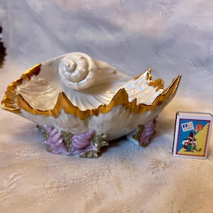 May include: A decorative, shell-shaped porcelain dish with a pearlescent interior and gold-painted edges. The dish is supported by pink and green coral-like accents. A matchbox with a colorful illustration is next to the dish.
