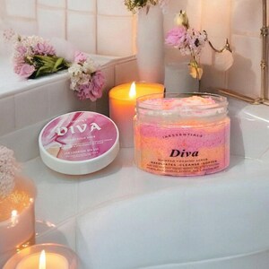 Puede incluir: Un tarro de exfoliante espumoso batido Diva con capas rosas, naranjas y blancas, y un recipiente de exfoliante de azúcar Diva a juego. Los productos se exhiben con velas encendidas y flores.
