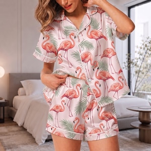 Puede incluir: Un conjunto de pijama de dos piezas que incluye una camisa de manga corta con botones y pantalones cortos a juego. La tela es de color rosa claro y presenta un estampado repetitivo de flamencos rosas y hojas de palma verdes.