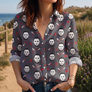 May include: A long-sleeve button-down shirt in a dark gray color featuring an all-over repeating pattern of white horror movie character masks and silver knives with red blood splatters. The shirt has a classic collar and buttoned cuffs.