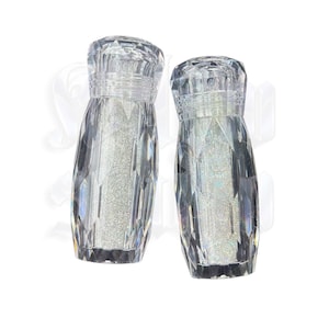 May include: Two clear, faceted salt and pepper shakers. The shakers have a diamond-like pattern and are filled with a shimmering, silver substance. The tops of the shakers are also faceted, and the bases are flat.