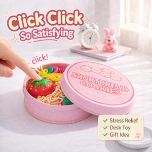 Puede incluir: Una lata rosa con la etiqueta "Shortbread Cookies" está abierta, revelando una colección de juguetes coloridos, incluyendo un tomate rojo y utensilios amarillos. El texto "Click Click So Satisfying" está encima de la lata. Un pequeño despertador y una figura de conejito rosa están en el fondo.