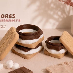 May include: S'mores-shaped containers in a stack and individually. The containers are brown, white, and tan, resembling graham crackers, chocolate, and marshmallow. The image includes the text "S'MORES Containers" and "Sweet storage for your little things."