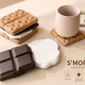 May include: A set of S'MORE COASTERS featuring individual pieces shaped like graham crackers, chocolate bars, and marshmallows. The coasters are brown, white, and dark brown, with a light-coloured mug resting on one square graham cracker coaster.
