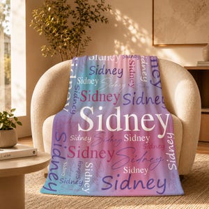 May include: A soft fleece blanket featuring a repeating pattern of the name Sidney in various fonts, sizes, and orientations. The background displays a gradient of blue, purple, and pink hues, with the name Sidney printed in white, purple, and pink.