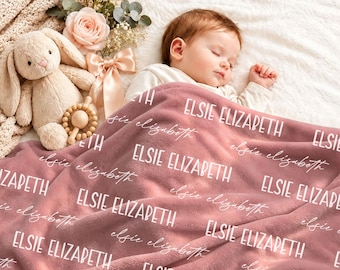 May include: A dusty rose plush blanket featuring the repeated white text "ELSIE ELIZABETH" in both a bold sans-serif font and a cursive script. The blanket is draped over a sleeping infant, accompanied by a beige plush rabbit toy.
