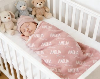 Könnte beinhalten: Ein schlafendes Baby in einem weißen Kinderbett, eingewickelt in eine rosa Decke. Die Decke ist mit den Namen "AMELIA" und "Thomas" in Weiß bedruckt. Das Baby trägt eine passende rosa Mütze. Kuscheltiere umgeben das Baby.