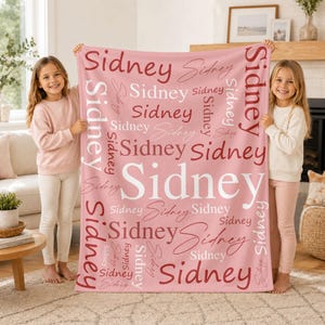 May include: A soft pink fleece blanket featuring the name Sidney printed repeatedly in various fonts, sizes, and shades of white, light pink, and dark red across the entire surface in a repeating pattern.