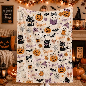 May include: A cream-colored fleece blanket featuring a repeating Halloween pattern of ghosts, black cats, and pumpkins. The design includes the name Siyana printed in purple and orange, alongside spiderwebs, bats, bows, and autumn leaves.