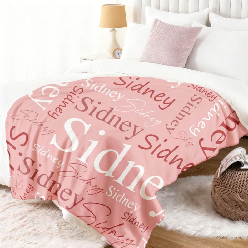 May include: A soft pink throw blanket featuring the name Sidney printed repeatedly in various fonts, sizes, and shades of pink and white. The text is arranged in a scattered, overlapping pattern across the entire surface of the fabric.