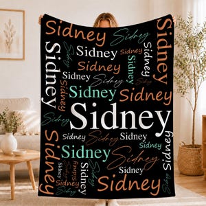 May include: A black rectangular throw blanket featuring the name Sidney printed repeatedly in various sizes, orientations, and fonts. The text appears in white, light blue, and peach colors, creating a layered, collage-style pattern.