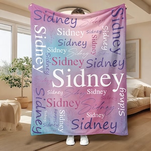 Könnte beinhalten: Eine personalisierte Decke mit dem Namen "Sidney" in verschiedenen Größen und Schriftarten wiederholt. Die Decke hat einen Farbverlaufshintergrund aus Blau-, Rosa- und Lilatönen. Das Wort "Sidney" ist in Weiß, Rosa und Lila geschrieben.