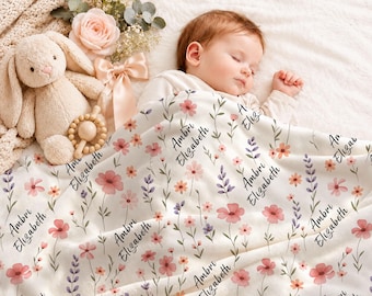 Könnte beinhalten: Eine weiße Babydecke mit einem wiederkehrenden Muster aus rosa und violetten Wildblumen und dem in schwarzer Schreibschrift gedruckten Namen Ambri Elizabeth. Die Decke liegt über einem schlafenden Säugling neben einem beigen Stoffhasen und einer Holzrassel.