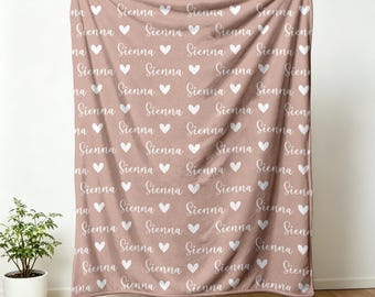 Könnte beinhalten: Eine altrosa Decke mit dem Namen "Sienna" in weißer Schreibschrift und weißen Herzmotiven. Diese weiche Decke ist ideal, um einem Schlaf- oder Wohnzimmer eine persönliche Note zu verleihen.