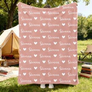 May include: A dusty rose blanket with a repeating pattern of the name "Sienna" and white hearts. The blanket is held up in front of a person, with a tent and outdoor setting in the background. The blanket is made of a soft material.