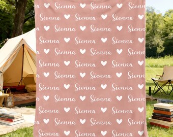 May include: A dusty rose blanket with a repeating pattern of the name "Sienna" and white hearts. The blanket is held up in front of a person, with a tent and outdoor setting in the background. The blanket is made of a soft material.