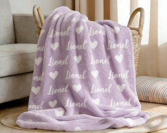 Könnte beinhalten: Eine fliederfarbene Fleece-Decke mit weißen Herzen und dem Namen "Lionel" im Druck. Die Decke liegt über einem Weidenkorb, ein gemütliches Wohnaccessoire.