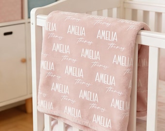 May include: A soft, dusty pink baby blanket draped over a white crib rail. The blanket features a repeating pattern of the name Amelia in a bold, white sans-serif font, alternating with the name Thomas in a white, cursive script font.
