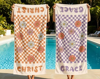 Könnte beinhalten: Zwei rechteckige Strandtücher mit wellenförmigem Schachbrettmuster und lächelnden Gänseblümchen. Ein Handtuch hat orangefarbene Akzente mit dem Text CHRIST, das andere violette Akzente mit dem Text GRACE. Beide Tücher werden aufrecht gehalten.