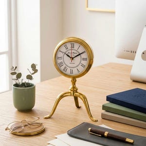 May include: A gold-colored desk clock with a white face and black Roman numerals. The clock is supported by a tripod base. The clock face reads "James Madison Oxford". Other items on the desk include a small plant, glasses, and a pen.