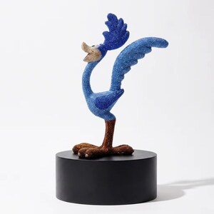 May include: A blue beaded Road Runner sculpture with outstretched wings and a raised head, mounted on a black cylindrical base. The bird's beak is tan, and its feet are brown. The sculpture is set against a white background.