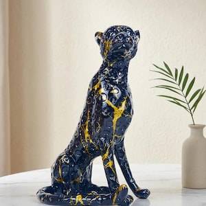 May include: A decorative ceramic leopard statue in a seated position. The statue is primarily navy blue with gold accents, featuring a textured surface. The leopard is sitting on a white marble table. A small vase with a green plant is in the background.
