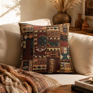 May include: Square decorative pillow with a patchwork design in shades of burgundy, teal, and beige. The pillow is placed on a beige sofa, with a patterned throw blanket and a stack of books visible in the foreground. The pillow's design resembles a vintage rug.