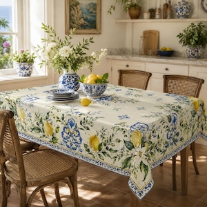 May include: A rectangular tablecloth featuring a cream background with a repeating pattern of yellow lemons, green leaves, and blue and white decorative floral motifs. The fabric drapes over a table set with a blue and white ceramic vase and bowl.