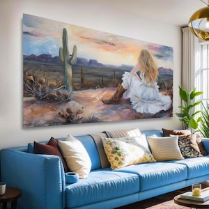 May include: A large rectangular canvas painting depicting a person with long blonde hair wearing a white dress and brown cowboy boots, sitting in a desert landscape with a tall saguaro cactus, distant mountains, and a soft sunset sky.