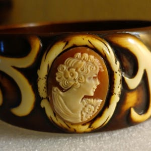 May include: Wide brown bangle bracelet with a cameo design. The bracelet features a central oval cameo of a woman's profile in cream, set against a brown background, and framed by cream-colored swirls.