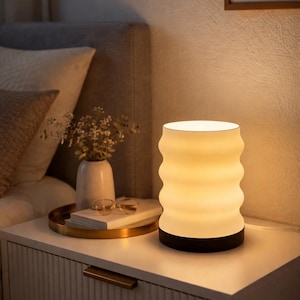 Könnte beinhalten: Eine weiß-schwarze Nachttischlampe mit wellenförmigem Design, die ein warmes Licht ausstrahlt. Die Lampe steht auf einem weißen Nachttisch, neben einem Tablett mit einer Vase, einer Brille und einem Buch. Der Raum hat eine neutrale Farbpalette.