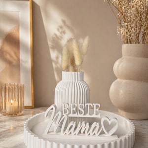 Puede incluir: Una bandeja decorativa blanca con borde acanalado sostiene un letrero blanco que dice "BESTE Mama" con un símbolo de corazón. Un jarrón blanco con flores secas y un segundo jarrón están en el fondo. También se ven una vela en un portavelas y una imagen enmarcada.