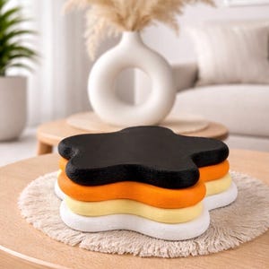 Könnte beinhalten: Ein Stapel blumenförmiger Untersetzer in Schwarz, Orange, Gelb und Weiß. Die Untersetzer liegen auf einer runden, beigefarbenen Untersetzer mit Fransen, die auf einem hellbraunen Holztisch platziert sind. Im Hintergrund eine weiße Vase mit getrockneten Pflanzen.