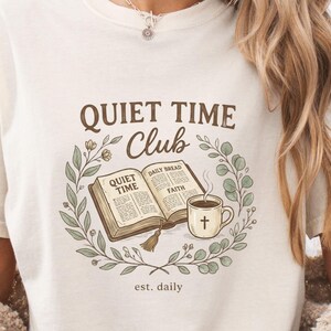 Puede incluir: Camiseta color crema con un gráfico de un libro abierto, una taza de café con una cruz y las palabras "Quiet Time Club" rodeadas por una corona. El texto "est. daily" está debajo. La camiseta se combina con pantalones cortos de mezclilla.