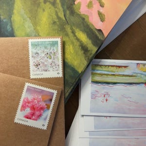 以下が含まれることがあります： Prunus Soine-yoshinoとPrunus Kanzanの桜の画像が描かれたカナダの切手が貼られた茶色のクラフト紙の封筒2枚と、ピンクと緑の色調の水彩風景画が描かれた長方形のカードの束。
