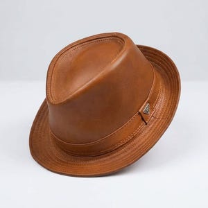 Può includere: Un cappello fedora in pelle color cuoio con corona strutturata e tesa larga. Il cappello presenta una fascia in pelle abbinata attorno alla base, rifinita con cuciture a vista e un piccolo emblema triangolare metallico applicato sul lato.