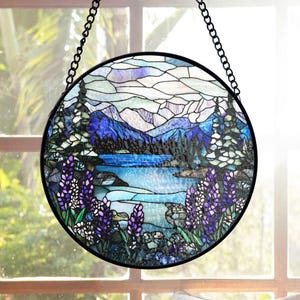 Könnte beinhalten: Runde Glasmalerei mit einer Bergsee-Szene. Das Kunstwerk zeigt einen blauen See, lila Wildblumen und immergrüne Bäume. Das Panel ist schwarz gerahmt und hängt an einer schwarzen Kette.