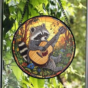 Op de afbeelding: Rond gebrandschilderd glazen raam met een wasbeer die gitaar speelt. De wasbeer is grijs en wit, met een zwart masker. De gitaar is bruin. De achtergrond bevat bomen, bloemen en muzieknoten. Een zwarte ketting is bevestigd om op te hangen.