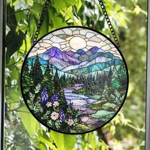 Könnte beinhalten: Ein rundes Buntglasfenster mit einer Berglandschaft mit Fluss, Bäumen und Blumen. Das Kunstwerk zeigt eine Sonne und ist mit einem schwarzen Metallrahmen und einer Kette zum Aufhängen versehen.