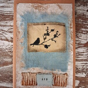 Op de afbeelding: Handgemaakte wenskaart met een vogel silhouet op een tak. De kaart heeft een textuurontwerp met een lichtblauw stoffen vierkant, een beige canvas en een kleine blauwe stoffen rechthoek met het woord "fly".