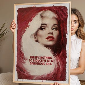 May include: A rectangular framed wall art print featuring a painted portrait of a blonde person with red lips against a deep red background. The text reads: THERE'S NOTHING SO SEDUCTIVE AS A DANGEROUS IDEA. The frame is light-coloured wood.