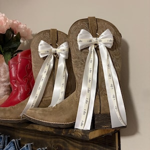 May include: Two brown suede cowboy boots with white ribbon bows that say "i do". The boots are on a wooden shelf with a red boot and pink flowers in the background. The ribbons hang down from the bows.