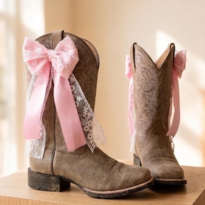 May include: A pair of brown cowboy boots with pink bows and lace accents. The boots have a classic western design with decorative stitching and a sturdy heel. The bows add a touch of femininity to the rugged footwear.