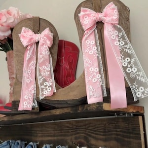 Puede incluir: Dos botas de vaquero marrones adornadas con lazos rosa bebé y cintas de encaje floral blanco. El texto "Baby pink + white floral lace" se muestra encima de las botas. Un par de botas de vaquero rojas son visibles detrás.