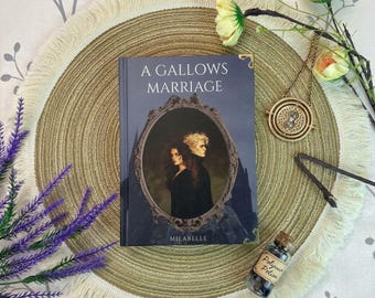 May include: A hardcover book titled A Gallows Marriage by Milabelle with a dark blue cover featuring an oval frame illustration of two people. Beside the book is a gold-toned circular pendant, a small glass bottle labeled Polyjuice Potion.