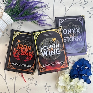 May include: Three hardcover books titled Iron Flame, Fourth Wing, and Onyx Storm by Rebecca Yarros. Each book features a black cover with gold-toned corner accents, intricate circular graphic designs, and dragon illustrations in red, orange, and purple.