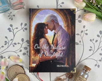 May include: A hardcover book titled On the Nature of Daylight by Ikorous, featuring an illustration of a couple embracing in an archway. The book is surrounded by two scented candles, two wooden coasters, a gold locket, and pink tulips.