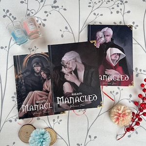 May include: Three hardcover books titled Manacled by SenLinYu, labeled Volume One, Volume Two, and Volume Three. Each cover features dark, illustrative artwork of two figures. Two small glass votive candles and decorative floral accents are present.