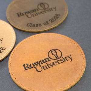 Könnte beinhalten: Drei runde Untersetzer aus Leder in Beige- und Brauntönen. Jeder Untersetzer zeigt das Logo und den Text "Rowan University". Ein Untersetzer ist mit "Class of 2026" markiert. Die Untersetzer haben eine genähte Kante.